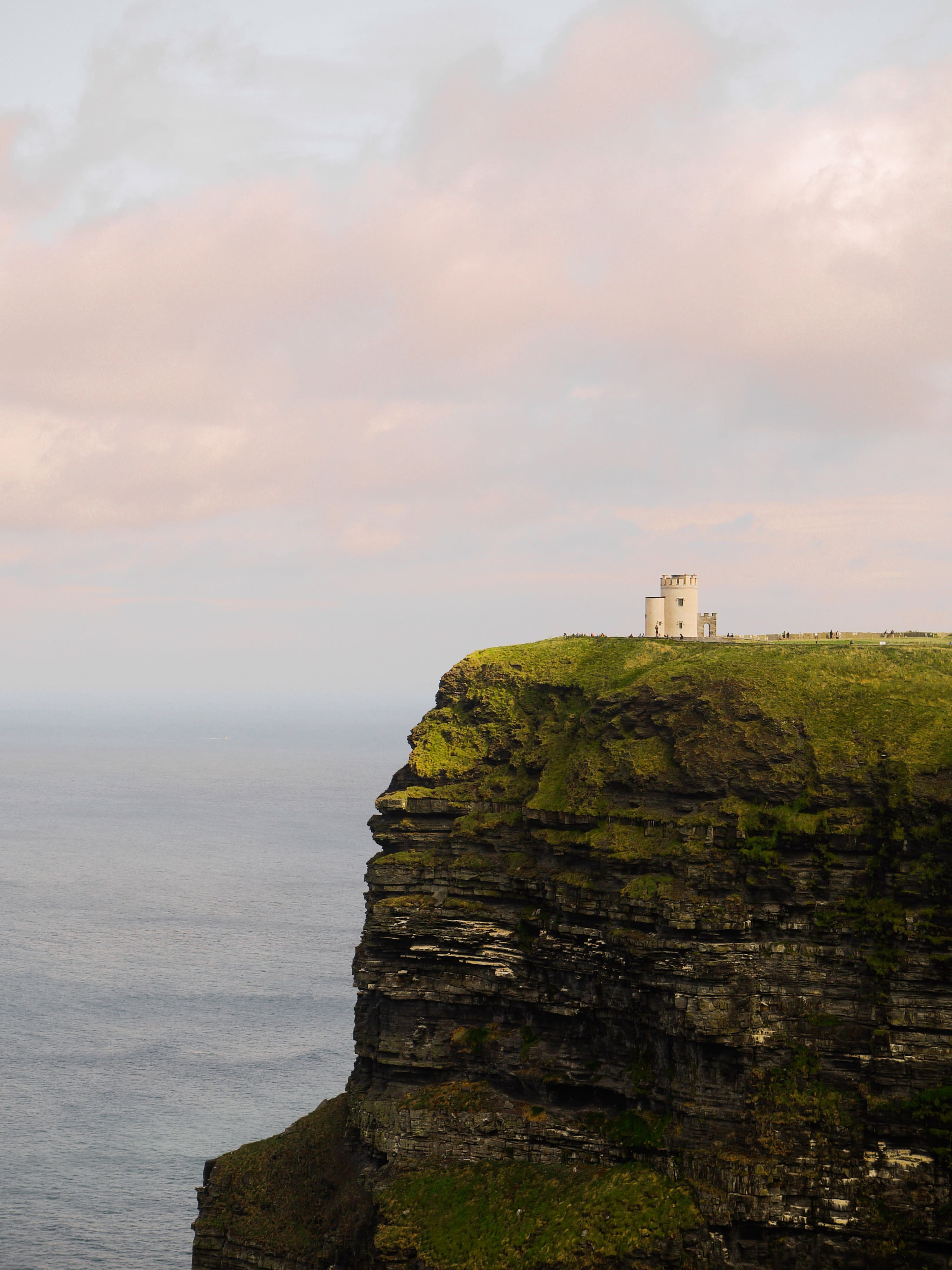 Green cliff with white building next to body of water with pink clouds in the sky during daytime