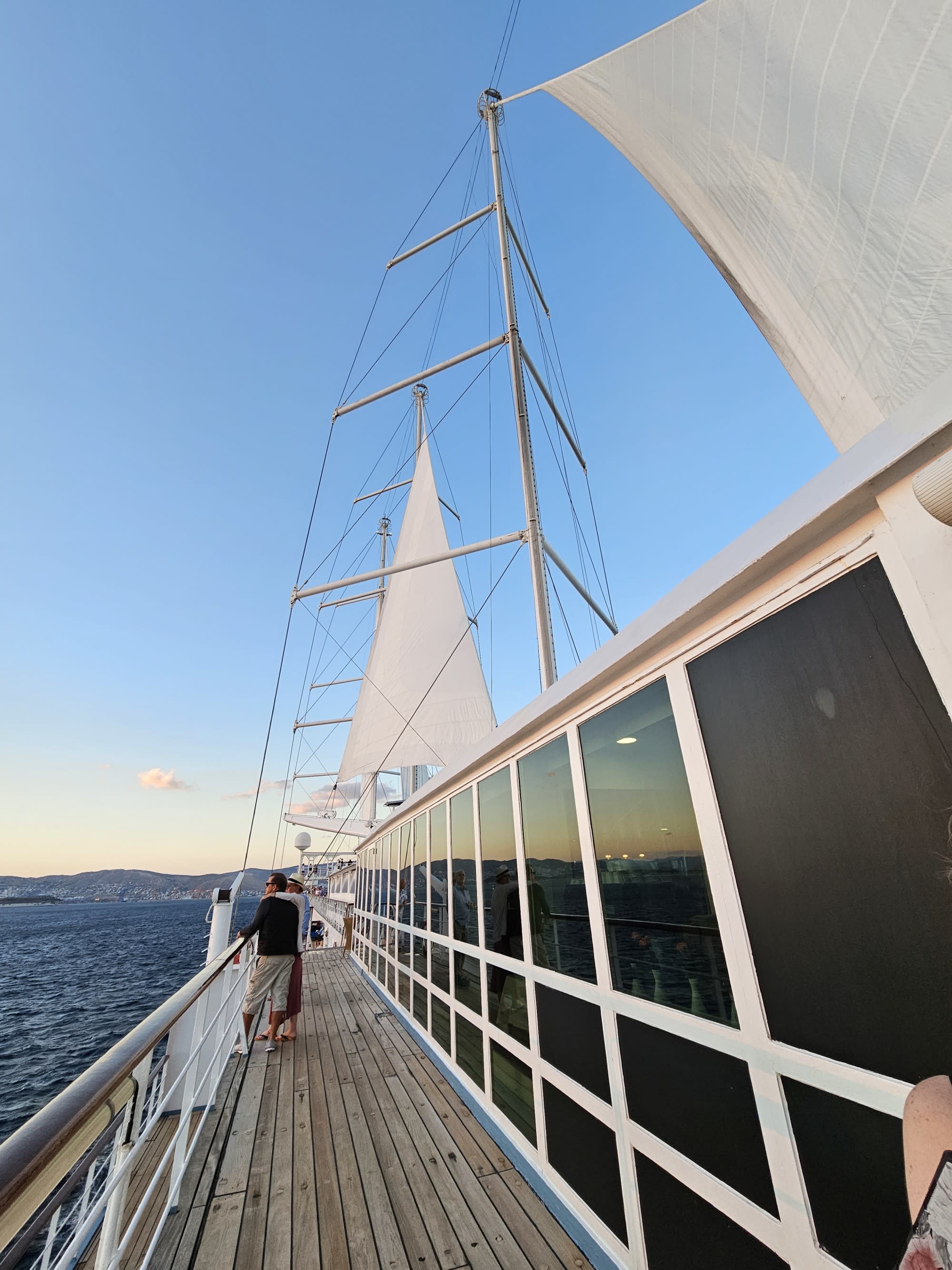 The image depicts a sunset view from the deck of a sailboat with sails unfurled and the horizon in the background.