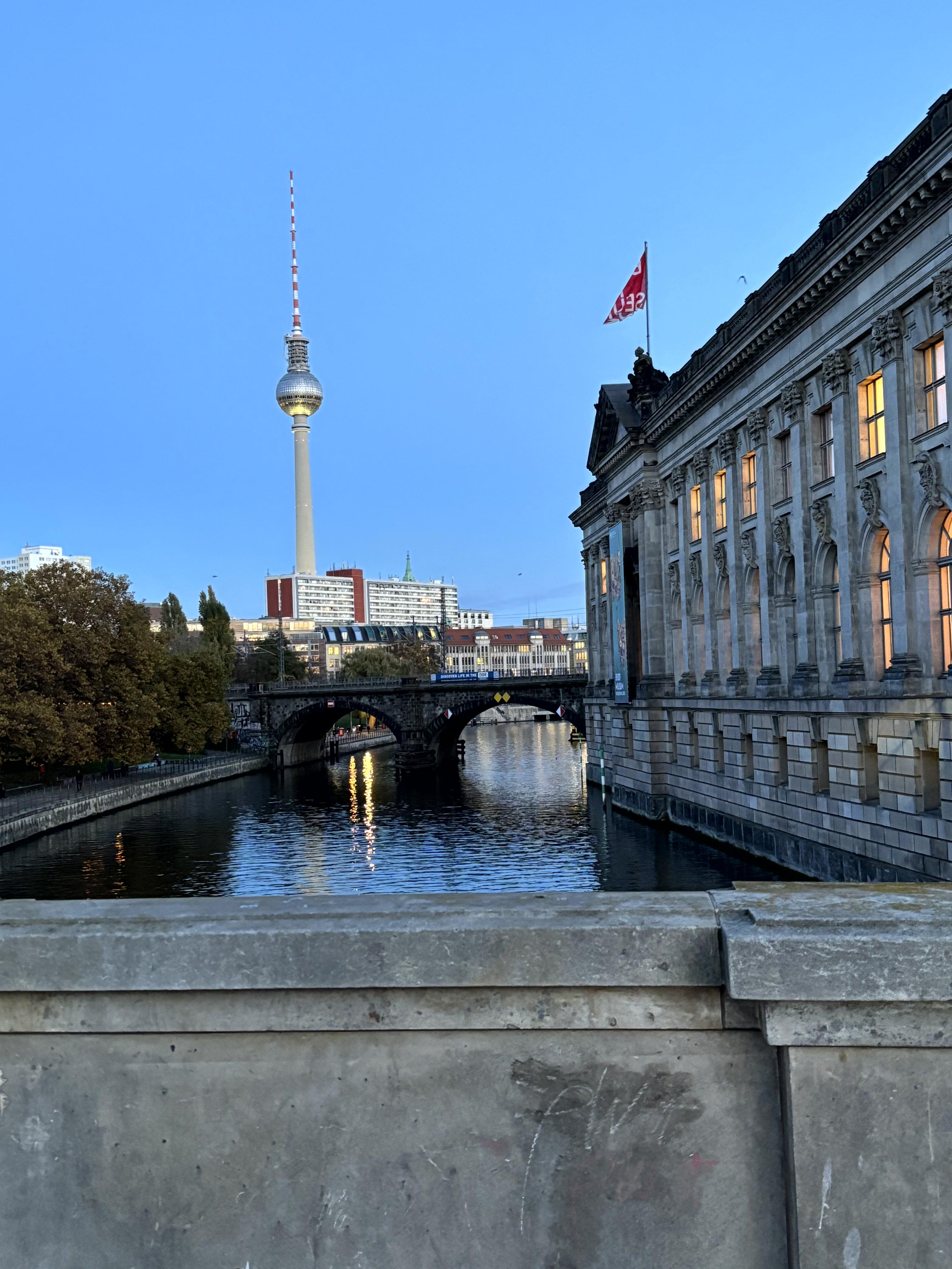 A stone building next to a river with the TV tower in the background, as seen from a concrete bridge in Berlin.