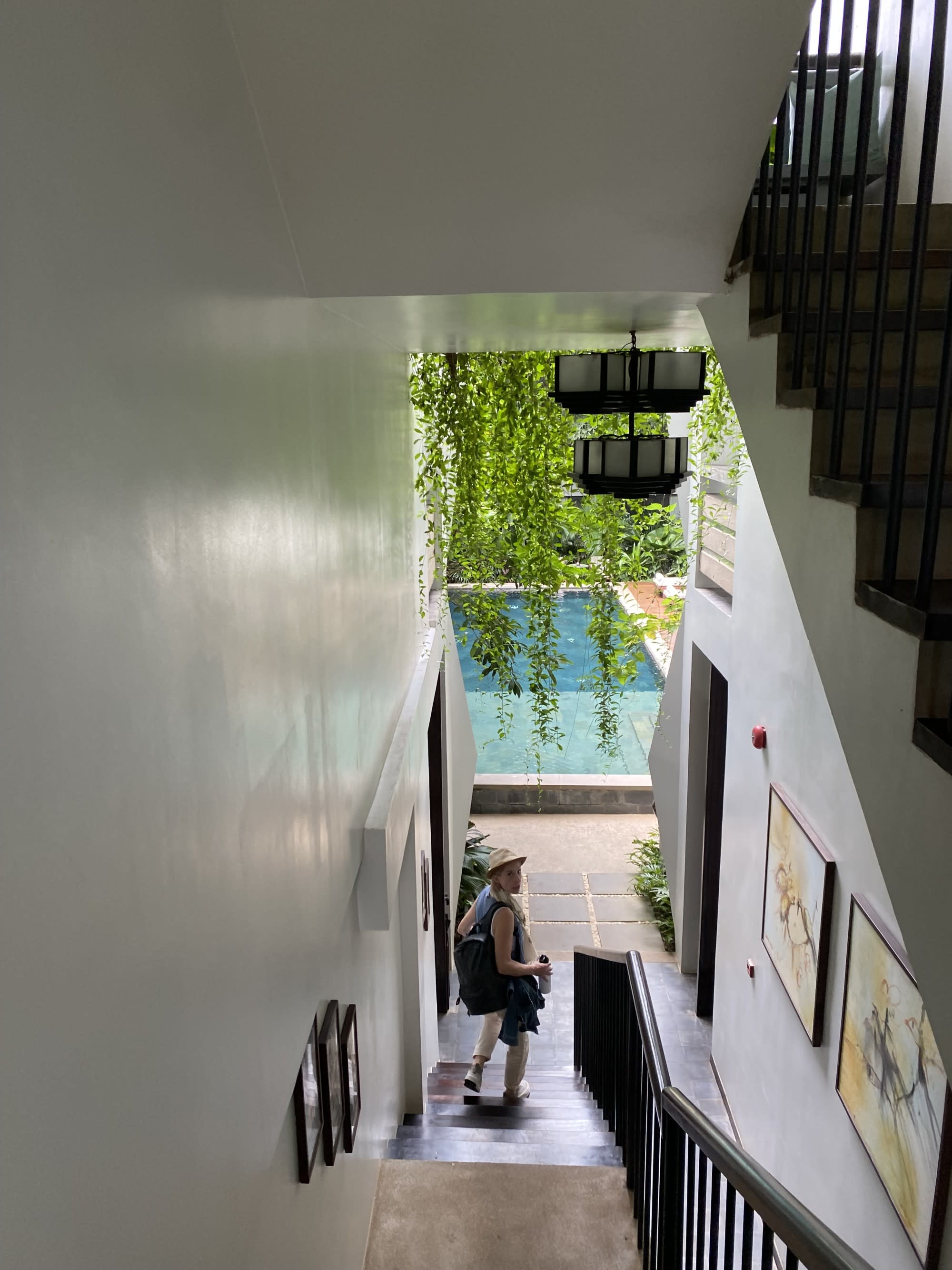 A woman standing at the foot of a staircase with large widows and an outdoor pool in the distance surrounded by foliage during the daytime.