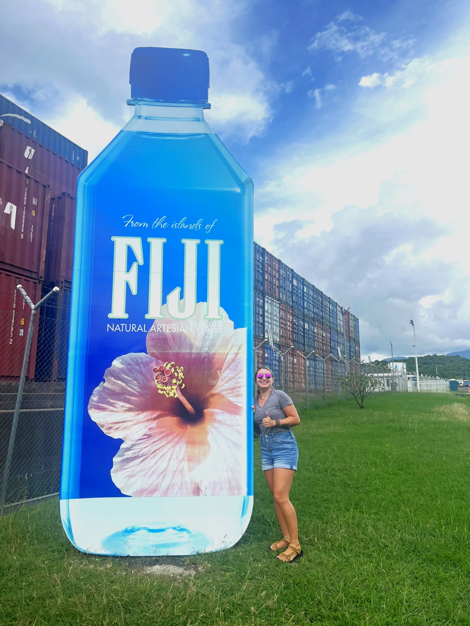 Danielle standing next to a large advertisement of a Fiji water bottle, with a clear sky and containers in the background.