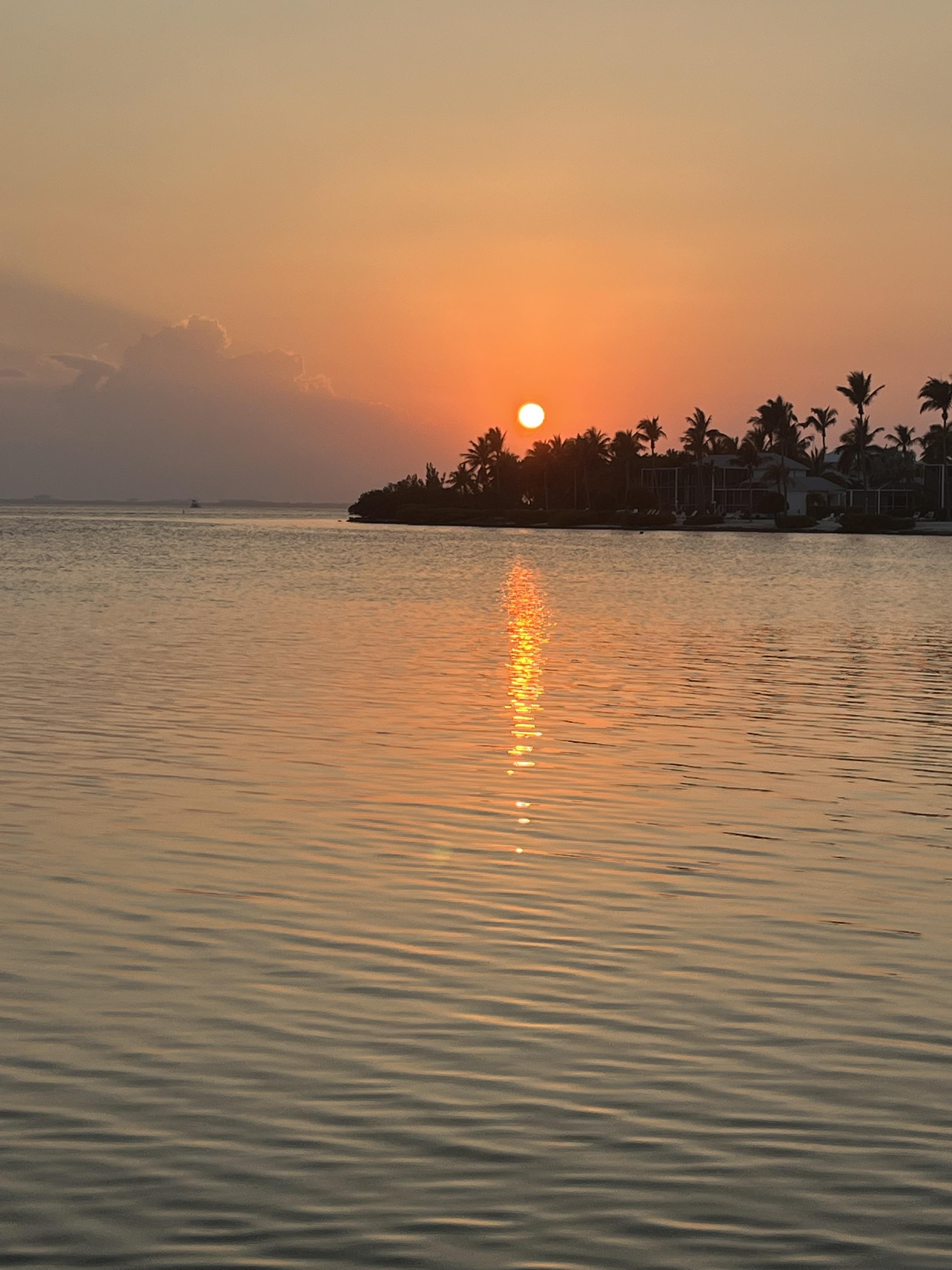 A bright orange sunset over Rum Point, a small peninsula with palm trees jutting out into the sea in the distance.