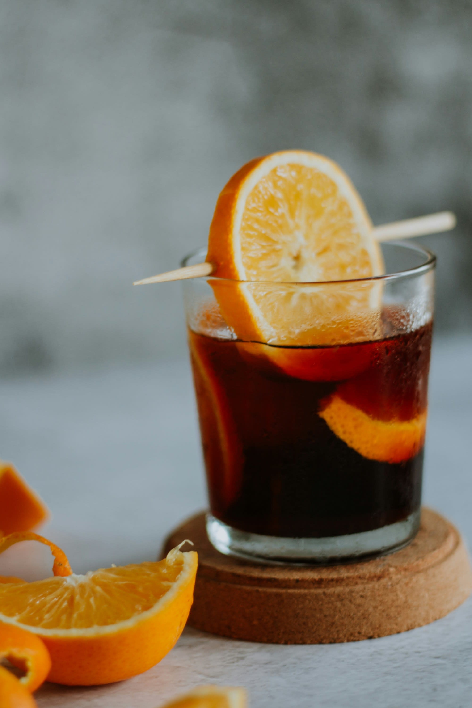This image depicts a cocktail with an orange slice and skewer poking through it resting on top of the beverage, which is also placed on top of a coaster. There are also orange slices on the bottom left side of the image.