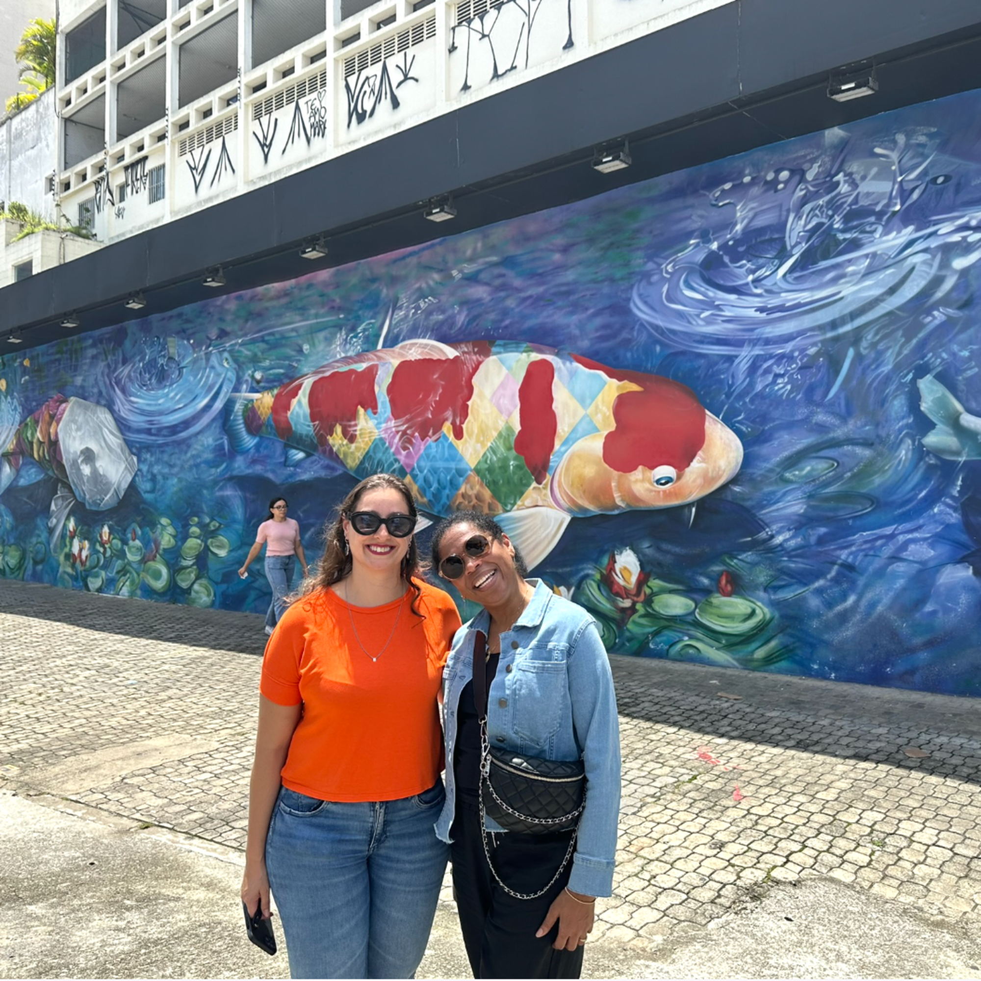 The image captures two people posing before a colorful mural depicting a large koi fish surrounded by marine life.