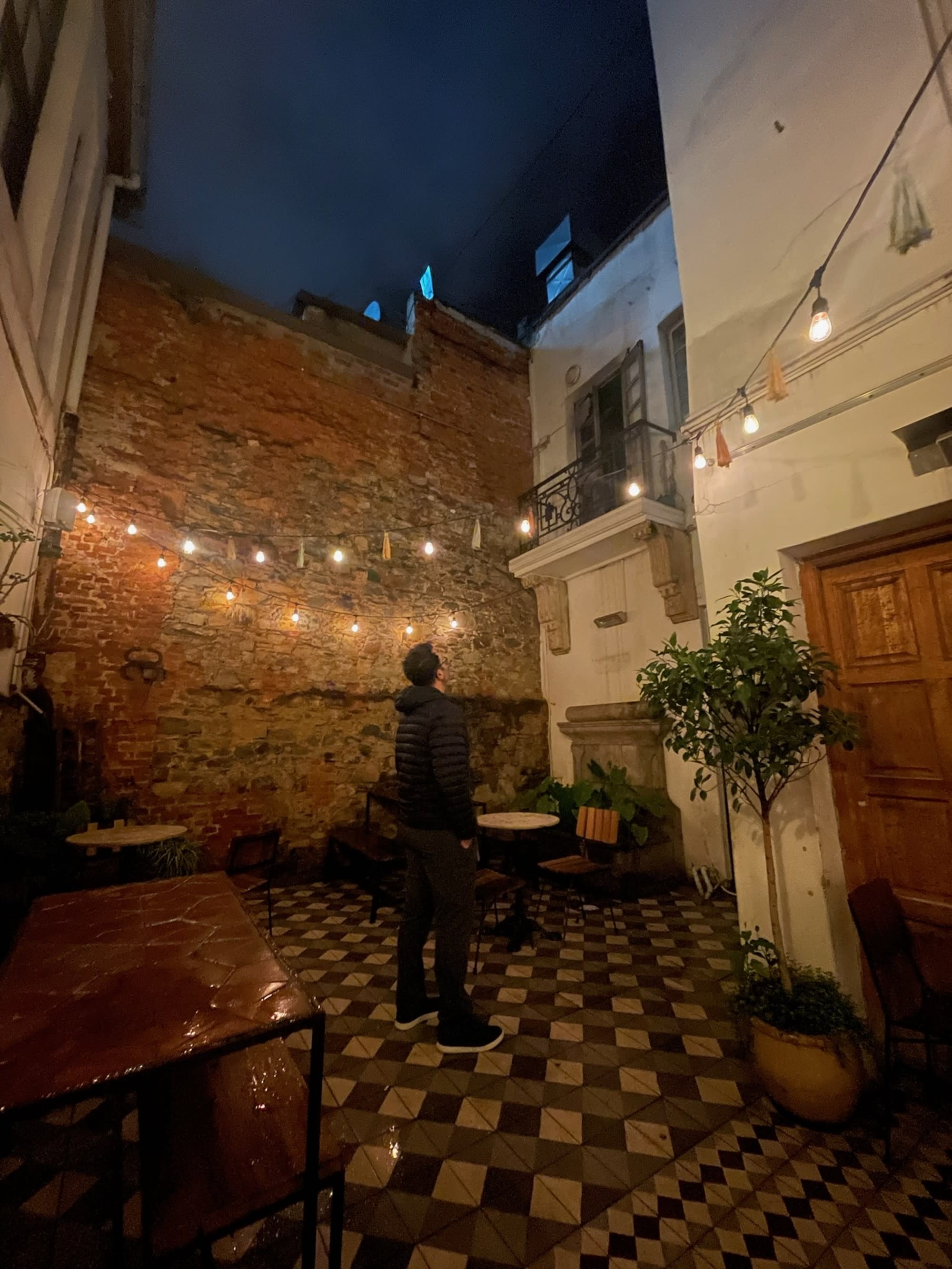 A person stands in a cozy, night-lit outdoor courtyard, surrounded by string lights and checkered tiles.