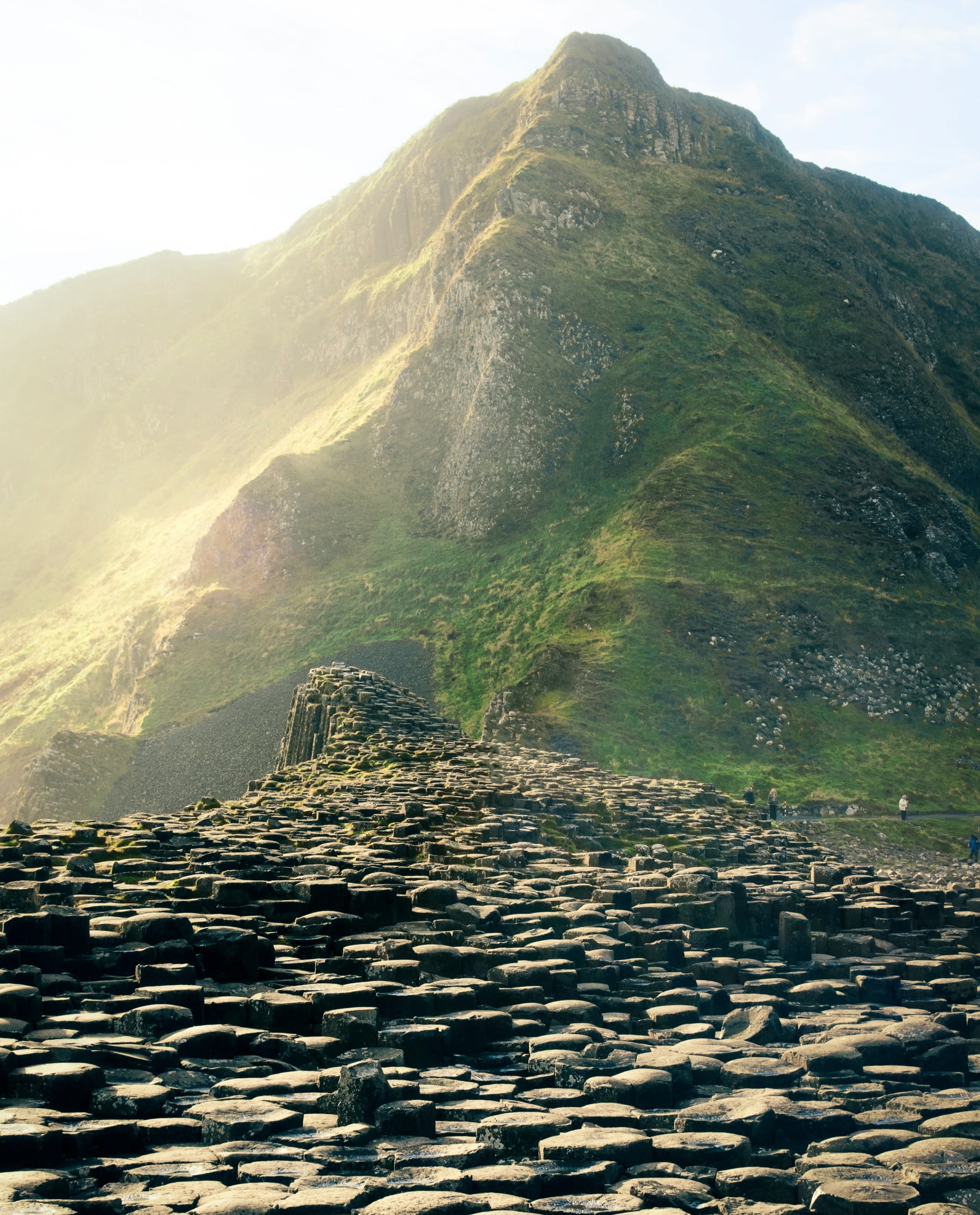 Giant's Causeway, one of the best things to do in Northern Ireland, with round rocks in a pathway leading to a green mountain on a misty, sunny day.