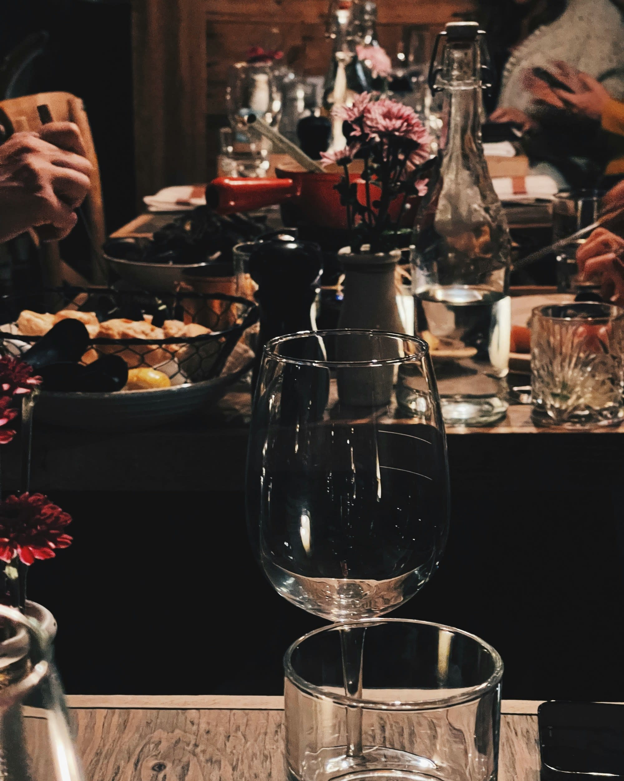 The image depicts a lively social dining scene with a table set with various glasses, bottles, and a vase with flowers.