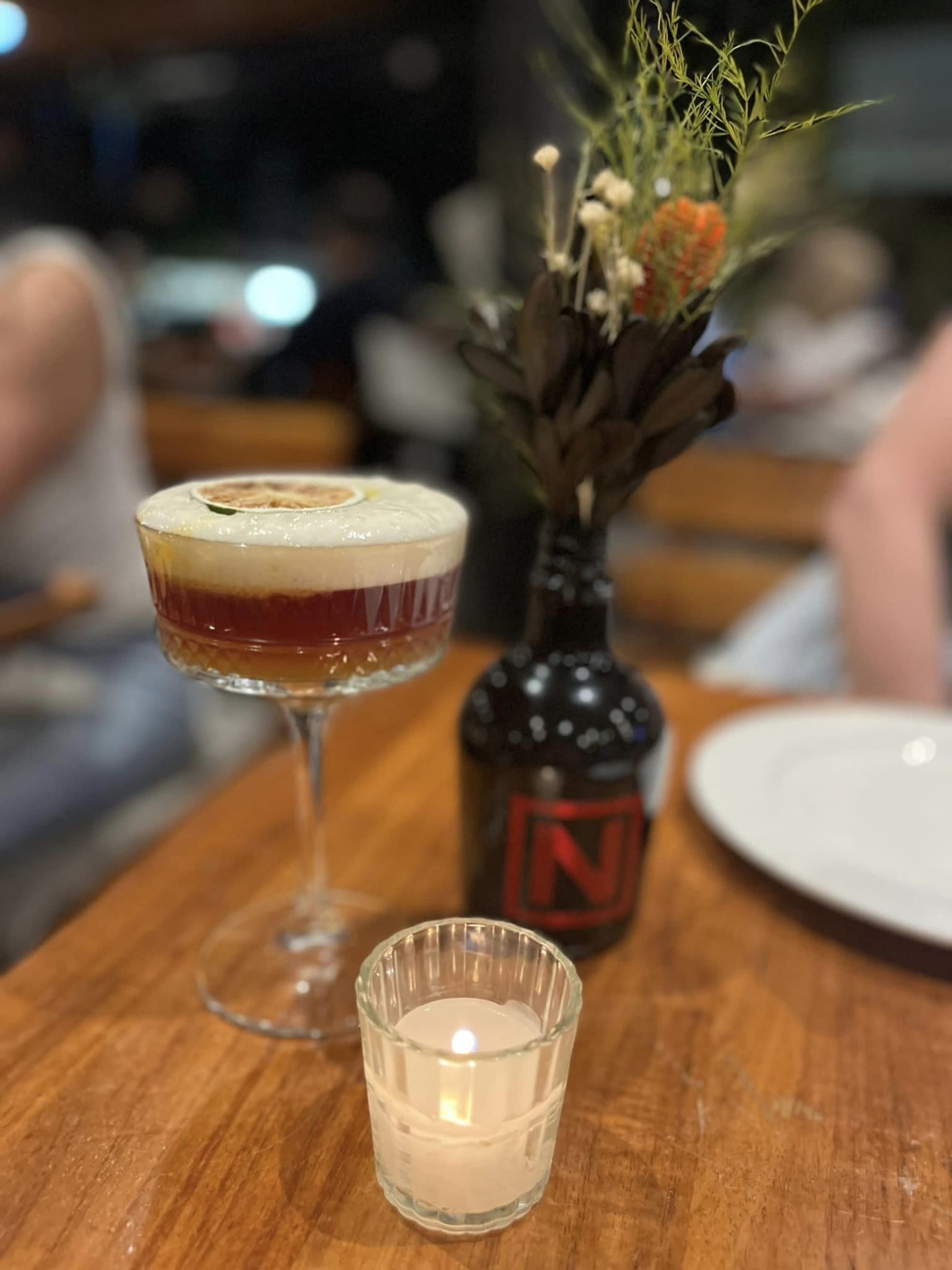 A sophisticated bar scene featuring a layered cocktail, fresh flowers, and the warm glow of candlelight.
