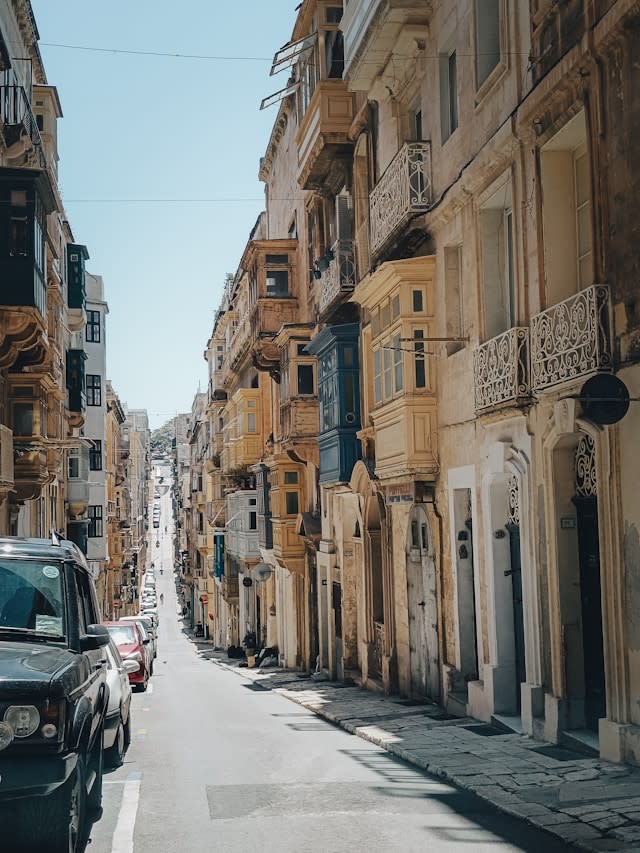 An image of a European city street with various stone balconies and detailing. There are cars parked along the left side of the street.