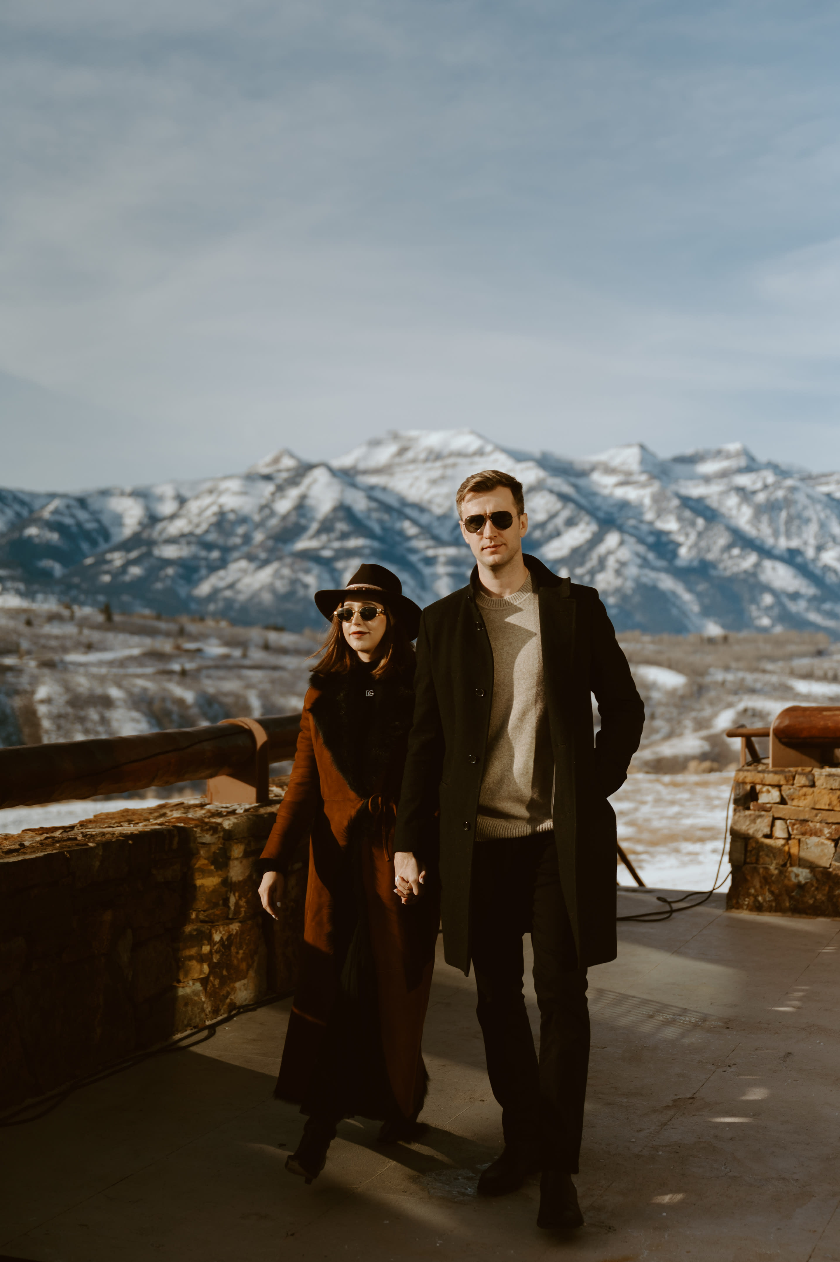 A couple dressed in swanky winter clothes, standing on an open balcony, posing in front of snow-covered mountains.