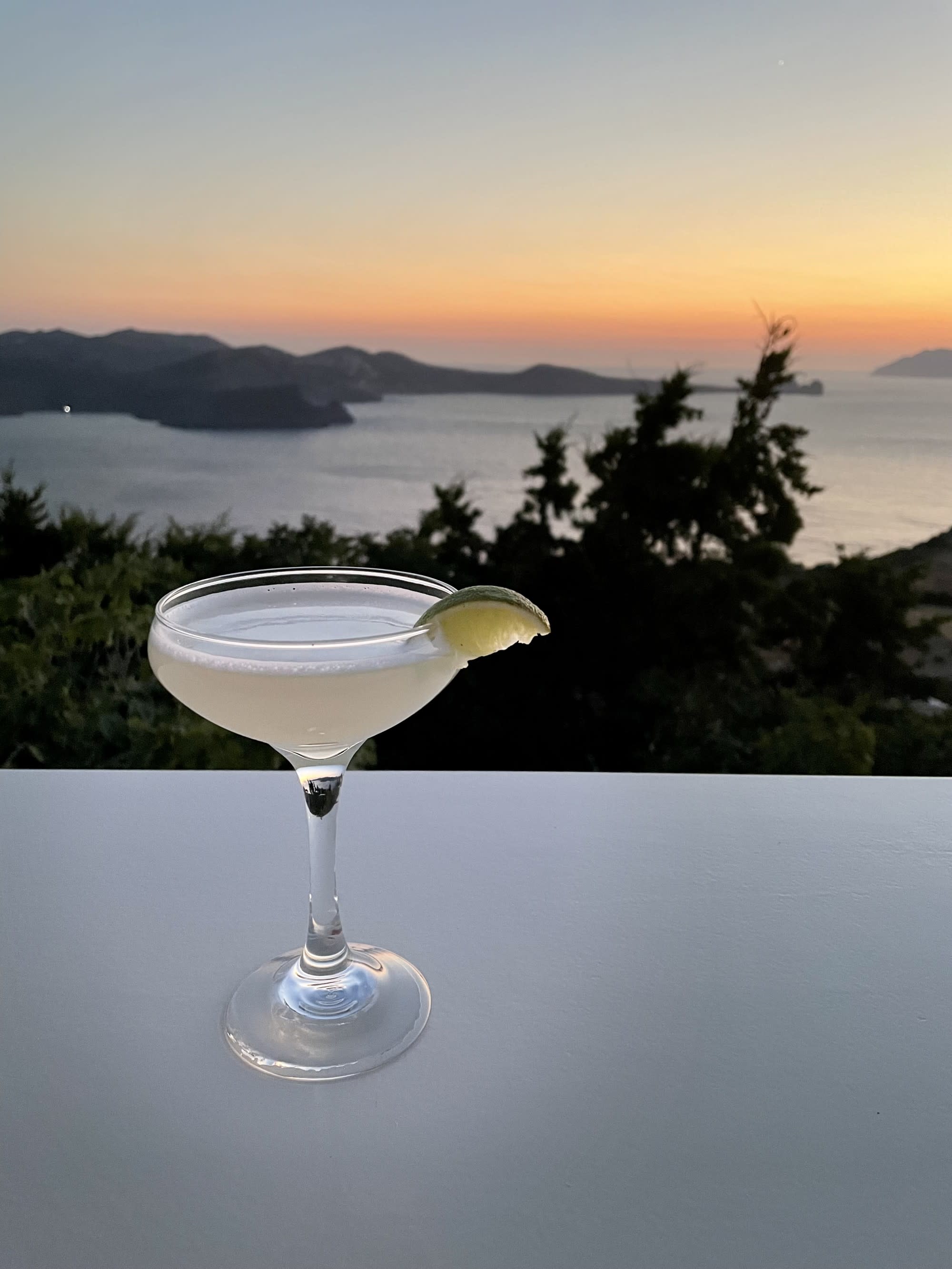 A close-up of an elegant cocktail set on a table, with a view of the sunset over the sea in the background.