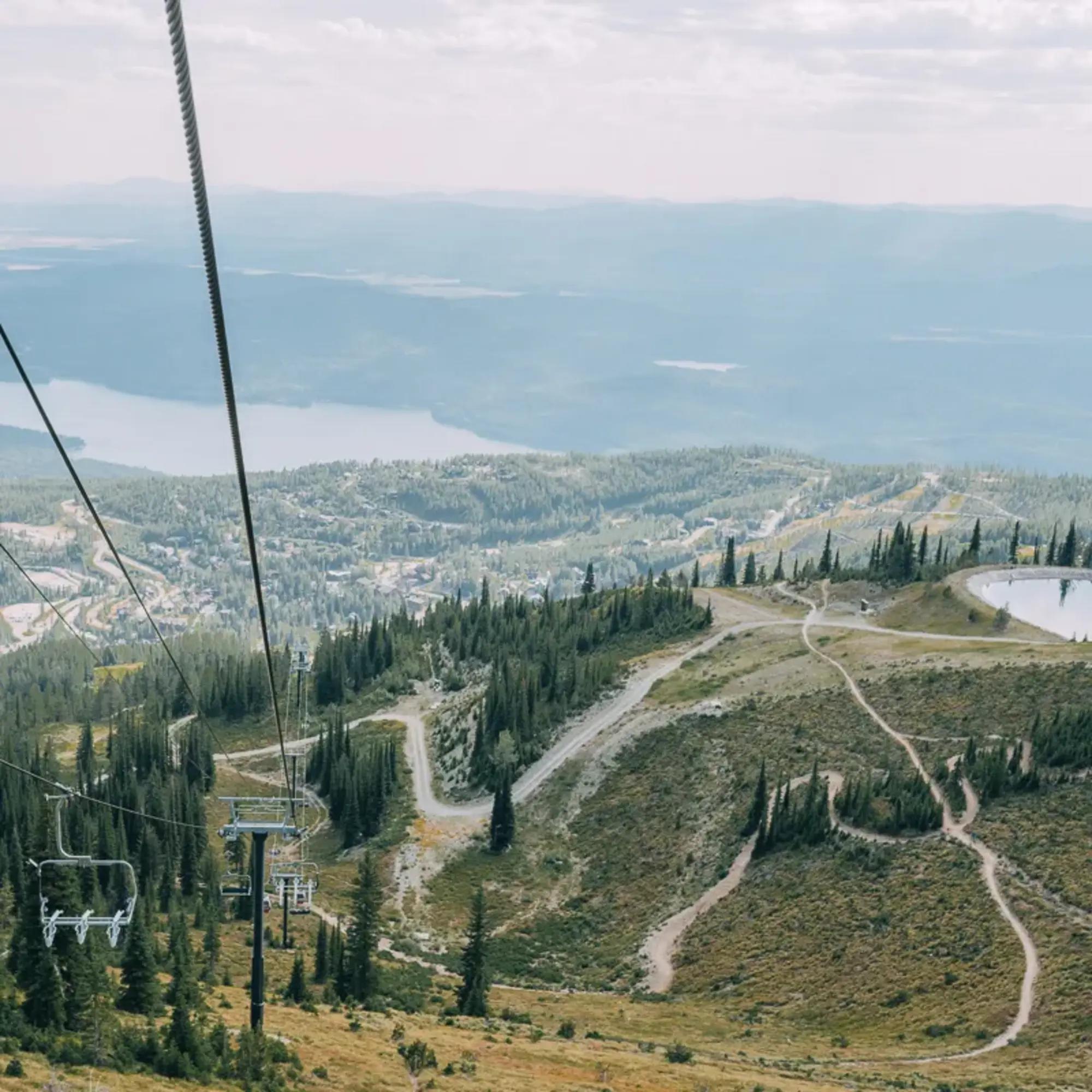 A picture of a ski hill, ski lift and pine trees with mountains and trails in the distance during the warmer seasons.