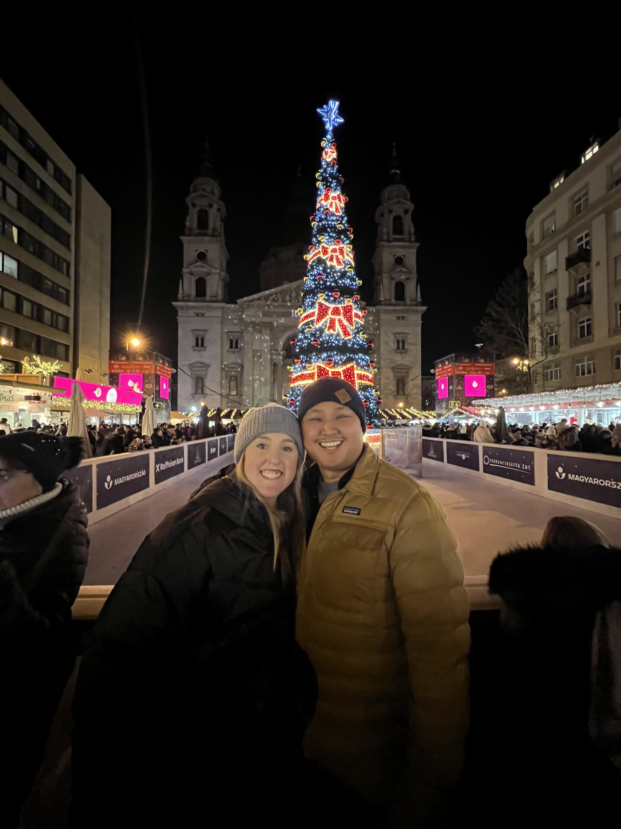 A couple posing for a photo in front of a lit up Christmas tree in a city square at night.