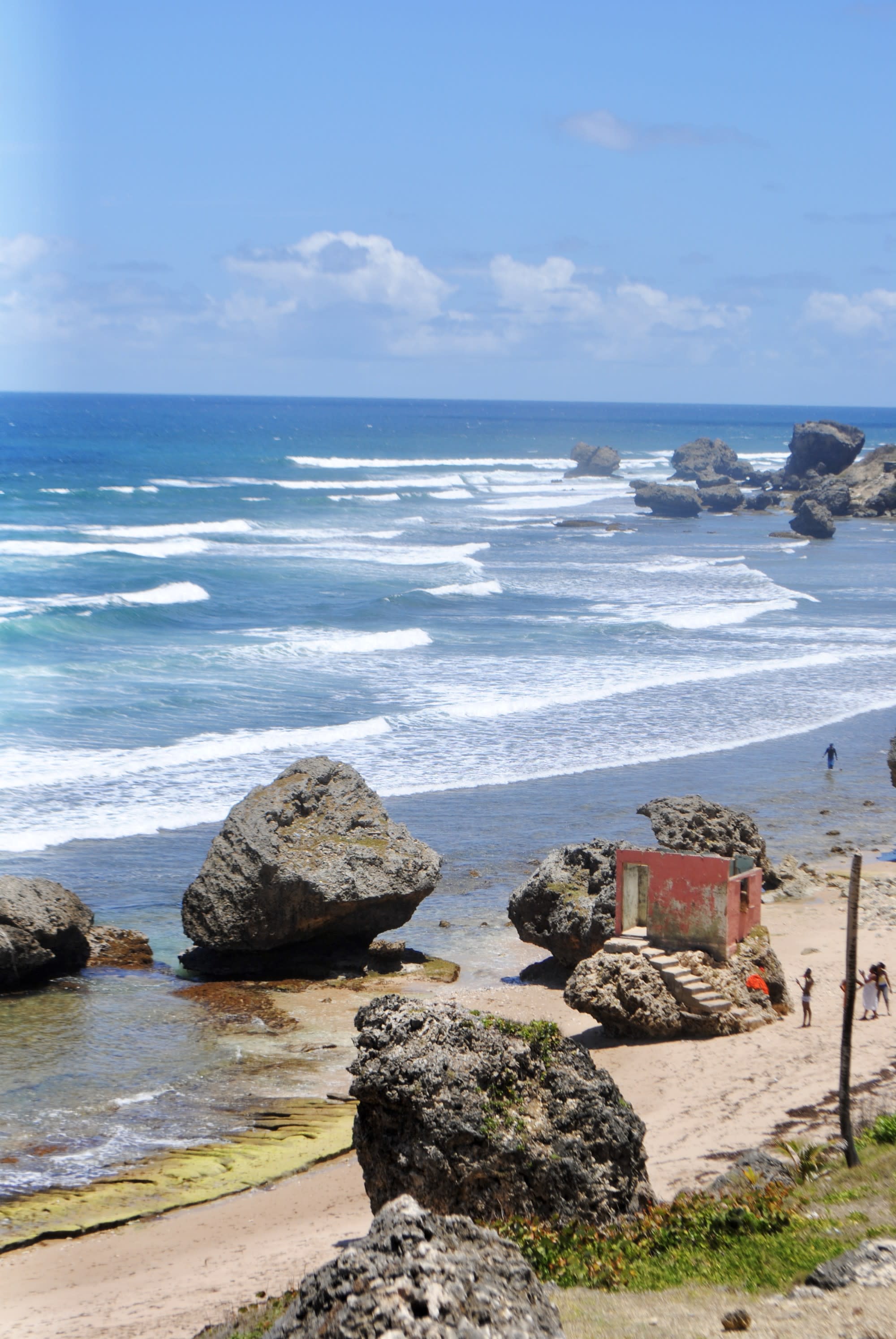 A view of the shore littered with big stone boulders, bright blue water and a blue sky.