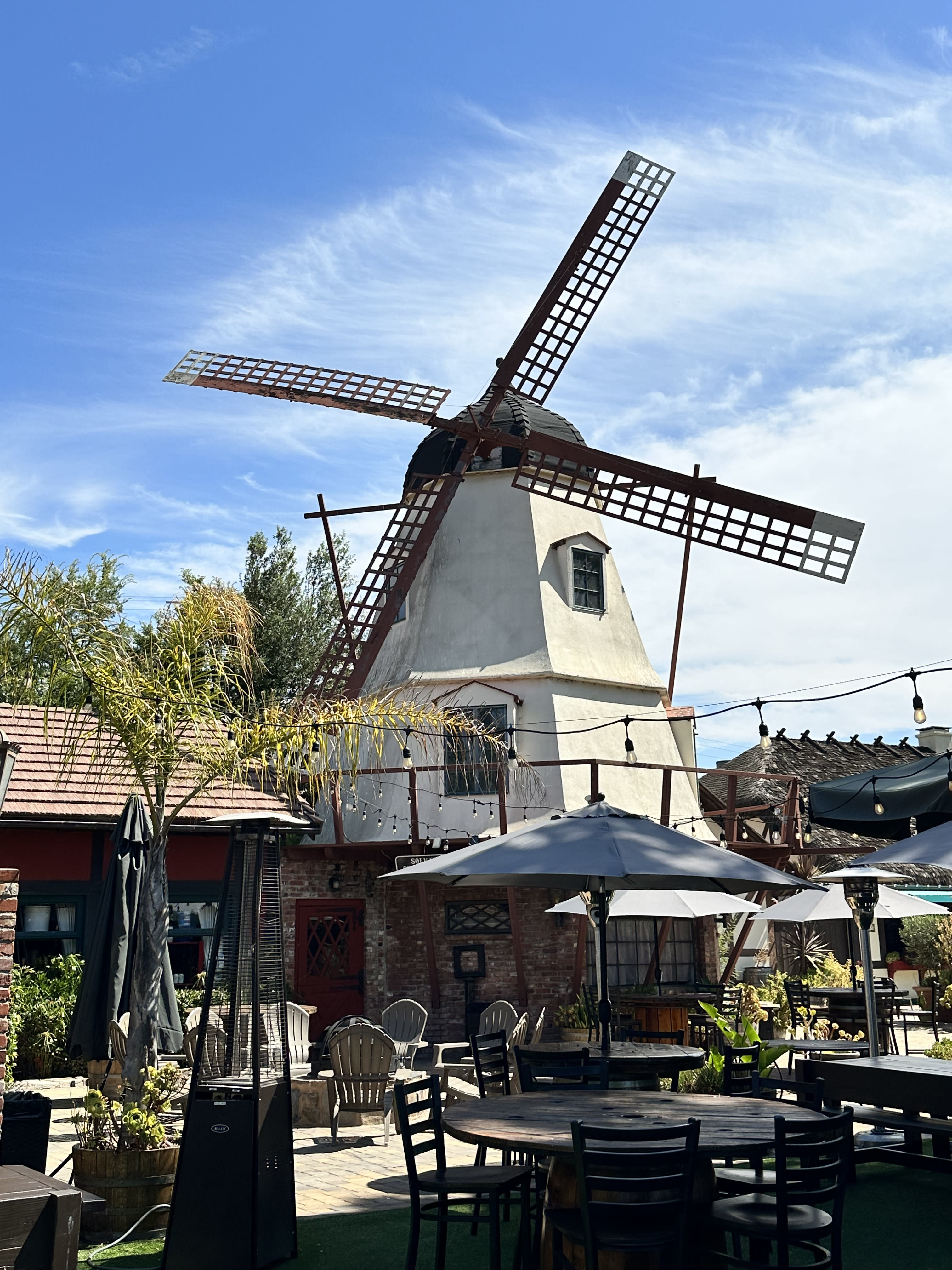 A windmill behind an outdoor patio with umbrellas, tables, chairs and string lights, as well as various plants.