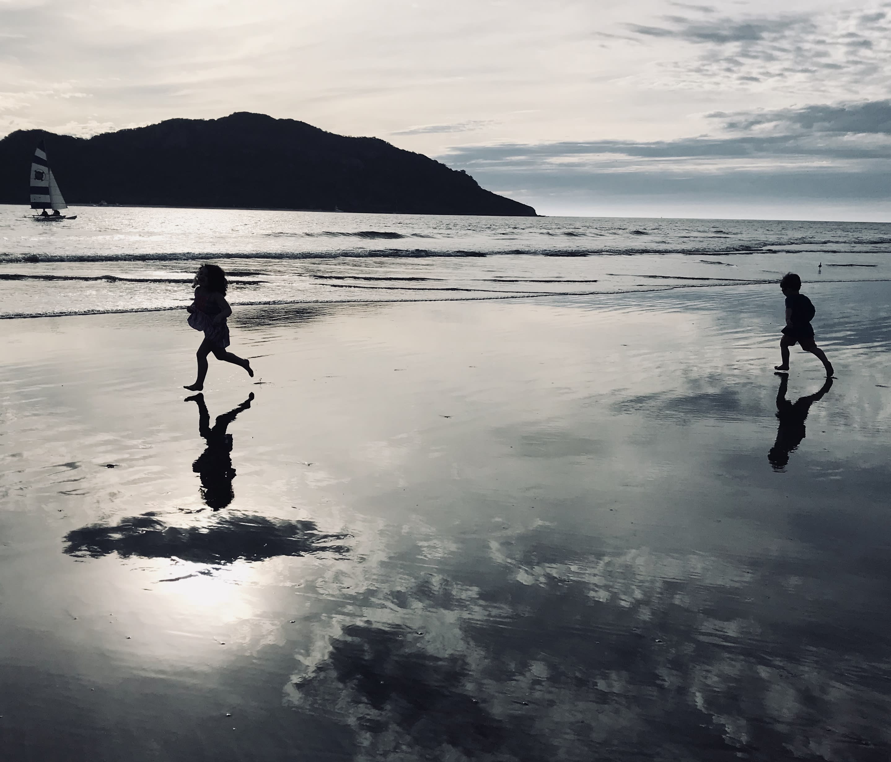 Two kids running on the beach with an ocean and a mountain in the background.