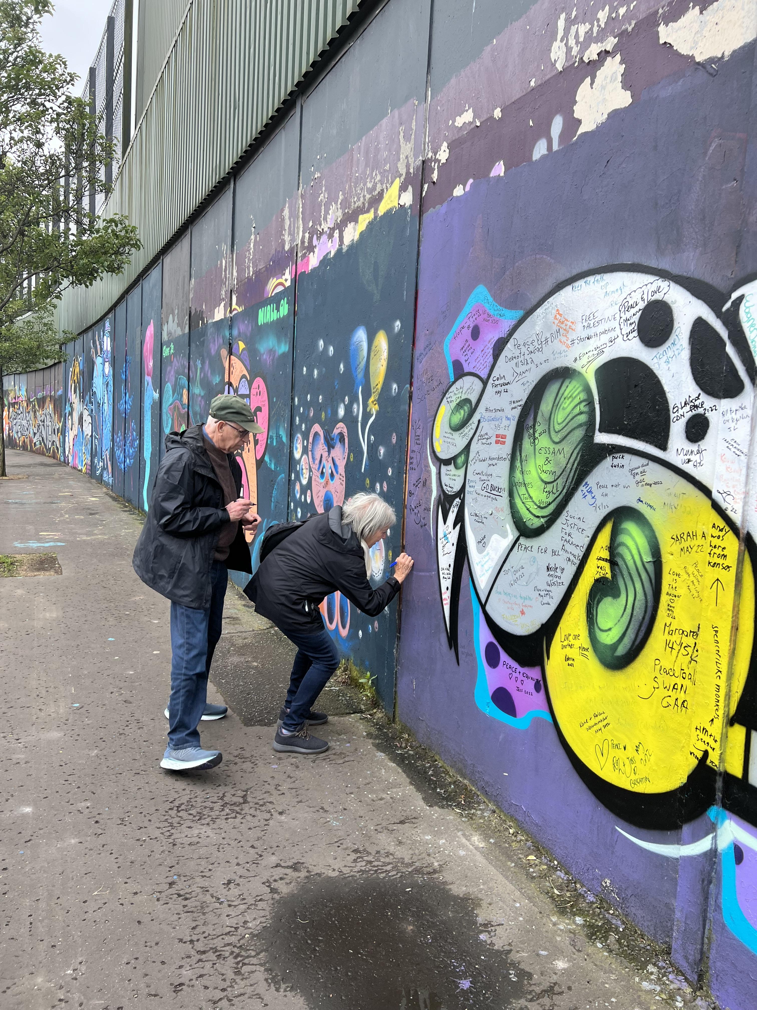 A woman writing on the colorful Belfast Peace Wall and a man standing behind her waiting to write.
