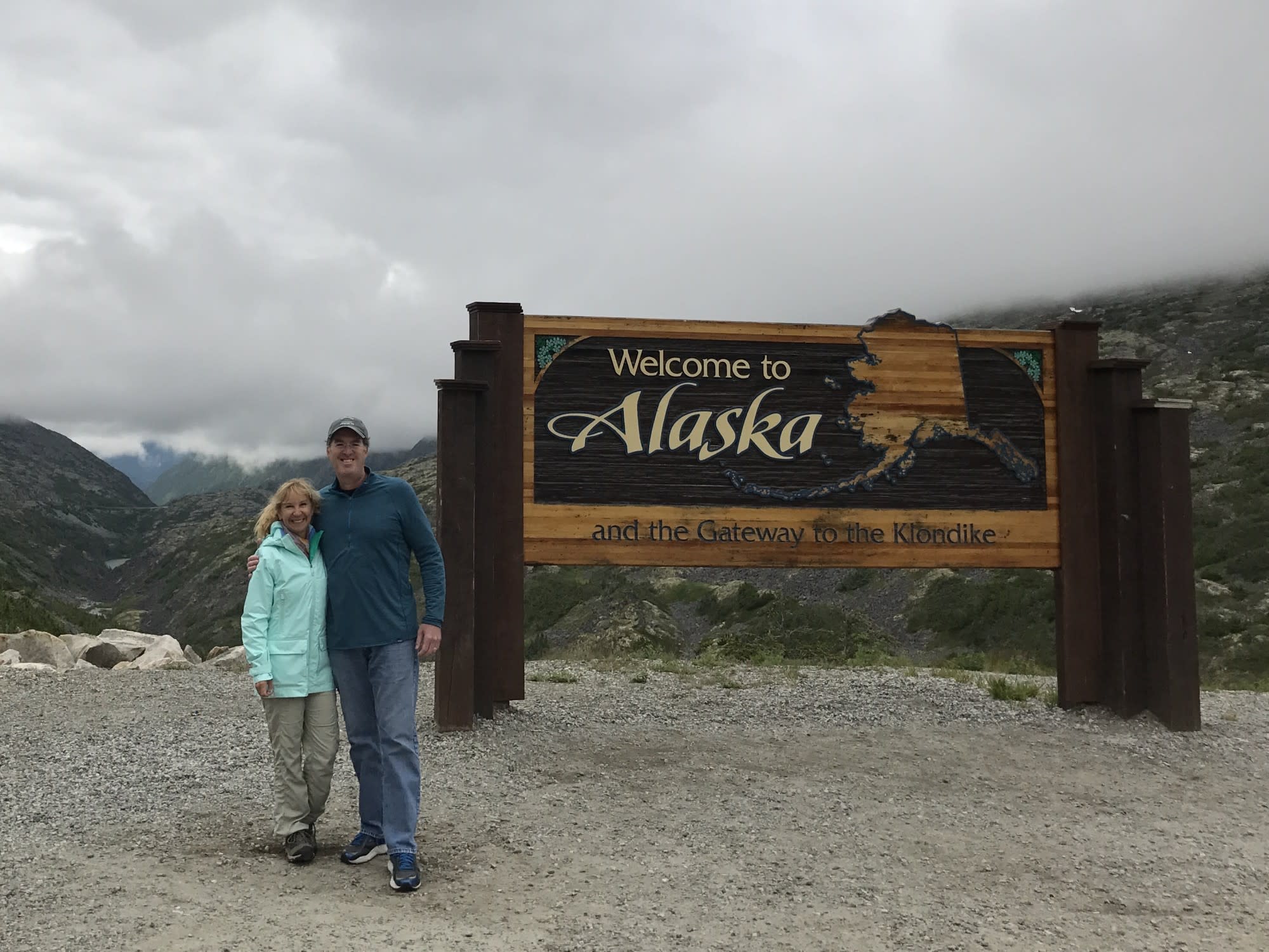 A couple standing in front of a "Welcome to Alaska" sign in front of a mountain landscape, during the best time to travel to Alaska.
